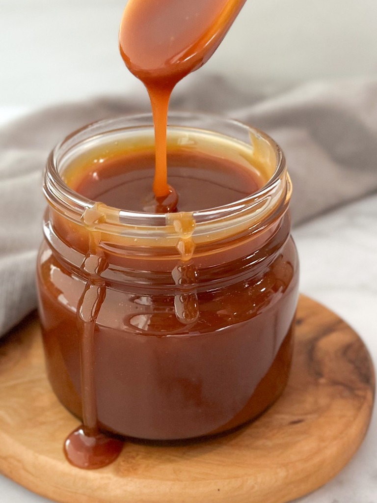 A rich salted caramel sauce is being poured into a glass jar, showcasing its smooth, glossy texture and inviting color.