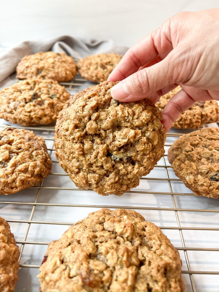 A hand grips a freshly baked gluten-free oatmeal raisin cookie resting on a cooling rack, emphasizing its inviting texture.