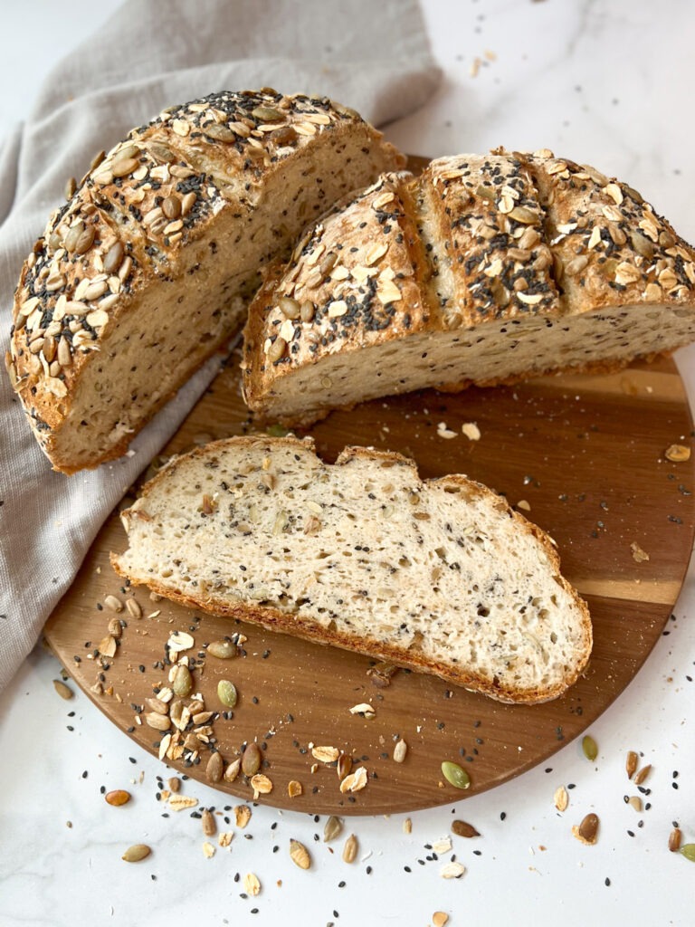 A loaf of gluten-free seeded honey oat boule is displayed on a cutting board, highlighting its crunchy seeds and nuts.